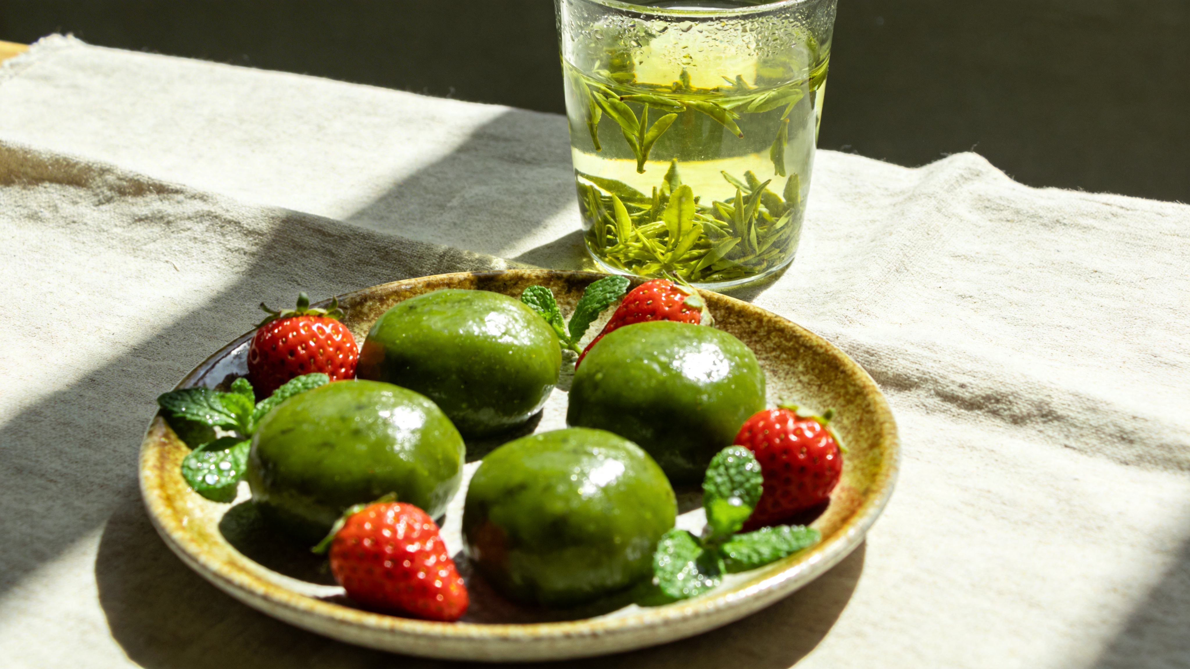 特写视角的春季时令美食静物摄影。画面中央是一个古朴的粗陶盘子，盘中盛着几块刚出炉的青团，表皮油润翠绿，那是用艾草汁染成的春日颜色。青团旁边散落着几颗鲜红的草莓和一把带露水的嫩薄荷作为点缀。背景是一块亚麻质地的餐布，上面放着一杯清透的玻璃杯，杯壁挂着水珠，里面泡着明前的龙井茶叶，茶叶在水中舒展，茶汤呈嫩黄绿色。午后的阳光从左侧45度角柔和地洒下，在青团表面形成诱人的高光，并在餐布上投下淡淡的、边缘模糊的阴影，营造出一种宁静、治愈且充满生活气息的下午茶氛围。

高端美食商业摄影风格，追求极致的真实感和材质纹理。采用大光圈虚化背景，突出主体。光线柔和自然，模拟窗边自然光，色彩还原真实且略微提高饱和度以突出食物的新鲜感。画质细腻锐利，能看清青团表面的颗粒感和草莓的籽，整体色调清新明亮，给人以食欲和舒适感。